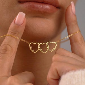 Può includere: Una collana dorata con tre ciondoli a forma di cuore interconnessi, ognuno con un nome: Nicole, Ethan e Paula. La collana è tenuta in mano, mostrando il suo design delicato e i dettagli personalizzati. La catena è sottile.