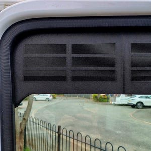 Könnte beinhalten: Nahaufnahme eines Fahrzeugfensterrahmens. Der Rahmen ist schwarz mit horizontalen Linien und einer weißen Oberseite. Das Fenster zeigt eine Straßenszene mit Autos, Bäumen und einem schwarzen Zaun.