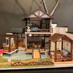 Puede incluir: Una maqueta en miniatura de una casa de estilo japonés con múltiples niveles, que presenta un interior y un exterior detallados. El modelo incluye un puente, cenadores y un pequeño jardín con plantas y flores en miniatura. La casa tiene un tejado marrón y paredes blancas.