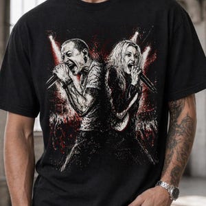 May include: A black short-sleeved t-shirt featuring a graphic print of two vocalists singing into microphones back-to-back. The illustration is rendered in black, white, and red, depicting a concert stage setting with spotlights and a crowd.