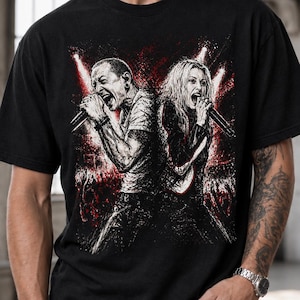 May include: A black short-sleeved t-shirt featuring a graphic print of two singers performing back-to-back on stage. The illustration uses black, white, and red ink to depict the performers holding microphones amidst bright stage lights and a crowd.