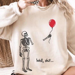 May include: A beige crewneck sweatshirt featuring a black line-art illustration of a skeleton looking up at a detached skeletal arm holding a red balloon. The text "Well, shit..." is printed in black below the arm, positioned to the right.