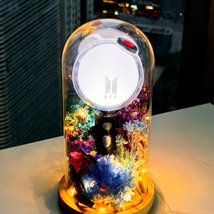 May include: A clear glass dome with a wooden base houses a BTS light stick surrounded by colorful dried flowers and fairy lights. The light stick has a white top with the BTS logo and a red button. The background shows a cityscape.