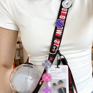Op de afbeelding: Zwarte en rode lanyard met witte tekst, met een heldere bolvormige lamp en verschillende kleurrijke bedels. De lanyard bevat ook een witte kaart met een wazige afbeelding. De lamp heeft een zwarte handgreep.