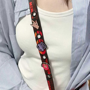 Peut inclure: Lanière noire avec texte rouge et diverses épingles. La lanière présente un motif répétitif de cercles et le texte "JUNGKOOK HITE". Les épingles comprennent un geste de la main, un logo et une épingle en forme de cœur. La lanière est portée sur un débardeur blanc et une chemise rayée.
