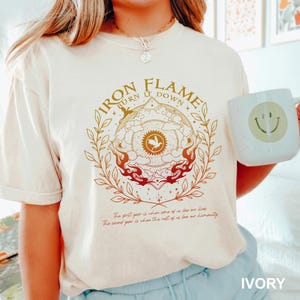 May include: Ivory-colored t-shirt with the text "IRON FLAME BURN IT DOWN" in a circular design with a bird and flames. The shirt also has text at the bottom. A white mug with a smiley face is held next to the shirt.