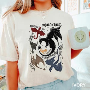 以下が含まれることがあります： 赤、黒、グレー、青のドラゴンテールが描かれたアイボリー色のTシャツ。「DRAGONTAILS」の文字がドラゴンのイラストの上にプリントされています。スマイリーフェイスの白いマグカップが画像に写っています。