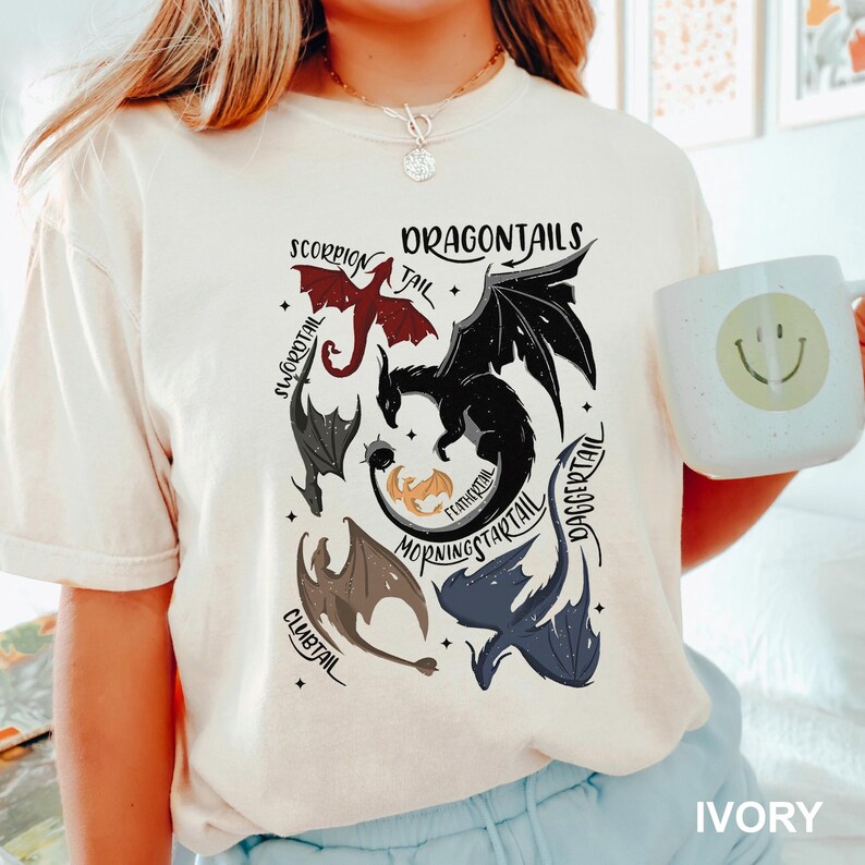 以下が含まれることがあります： 赤、黒、グレー、青のドラゴンテールが描かれたアイボリー色のTシャツ。「DRAGONTAILS」の文字がドラゴンのイラストの上にプリントされています。スマイリーフェイスの白いマグカップが画像に写っています。