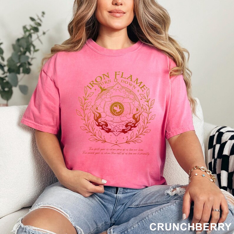 May include: A pink t-shirt with the text "IRON FLAME TURN IT DOWN" in a circular design with gold and red accents. The shirt has a crew neck and short sleeves. The bottom right corner of the shirt says "CRUNCHBERRY".