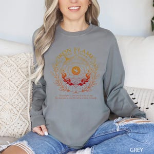 May include: A grey long-sleeve shirt with a circular graphic design in orange and red. The design includes the text "IRON FLAME TURNS IT DOWN" and other text below the graphic. The shirt is made of a soft material.