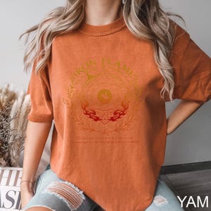 May include: Burnt orange t-shirt with a circular graphic featuring the text "IRON FLAME" and a decorative design in gold and red. The shirt has short sleeves and a relaxed fit, suitable for casual wear.