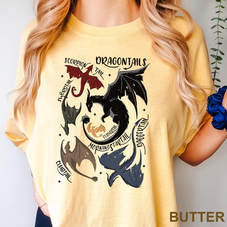 以下が含まれることがあります： ドラゴンのグラフィックが描かれた黄色のTシャツ。「Scorpion Tail」、「Swordtail」、「Daggertail」など、さまざまな種類のドラゴンがデザインされています。「DRAGONTAILS」の文字が上部にあります。「BUTTER」の文字が下部にあります。