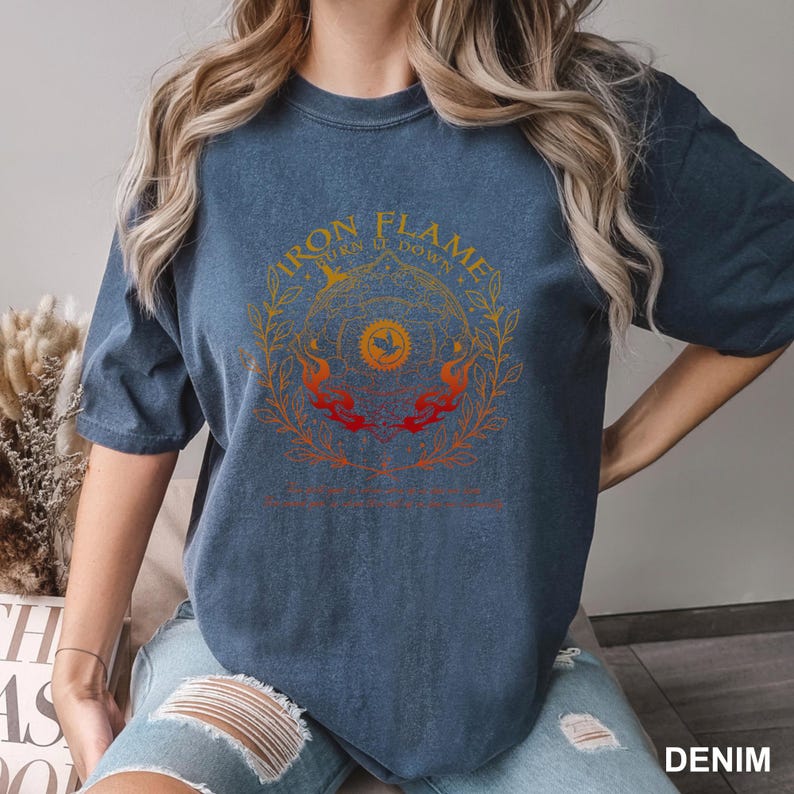 May include: A denim blue t-shirt featuring a circular graphic with the words "IRON FLAMES" and "TURN IT DOWN" in gold lettering. The graphic includes a sun, flames, and a laurel wreath design. The word "DENIM" is printed at the bottom.