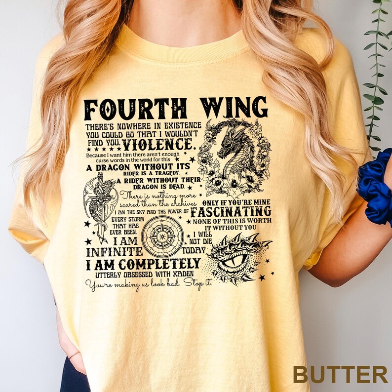 May include: Butter-colored t-shirt with black text and graphics. The text reads "FOURTH WING" and includes phrases like "A DRAGON WITHOUT ITS" and "I AM INFINITE." The design features a dragon, stars, and other decorative elements.