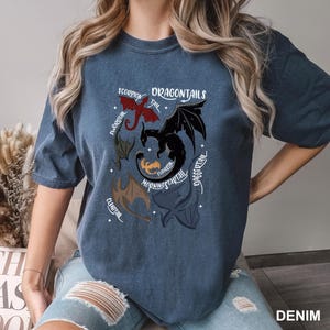 以下が含まれることがあります： デニムブルーのTシャツ。ドラゴン柄で、「DRAGONTAILS」の文字と、赤、黒、茶色のドラゴンのイラストが描かれています。右下には「DENIM」の文字がプリントされています。