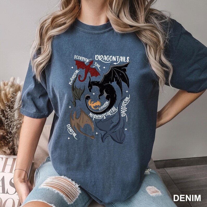 以下が含まれることがあります： デニムブルーのTシャツ。ドラゴン柄で、「DRAGONTAILS」の文字と、赤、黒、茶色のドラゴンのイラストが描かれています。右下には「DENIM」の文字がプリントされています。