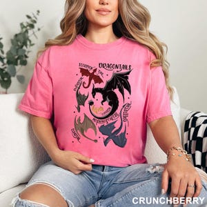 以下が含まれることがあります： ピンク色のTシャツ。ドラゴンをモチーフにしたデザインで、「Scorpion Tail」や「Daggertail」など、様々な種類の尻尾を持つドラゴンが描かれています。「DRAGONTAILS」の文字が上部にあります。Tシャツは単色のピンク色です。