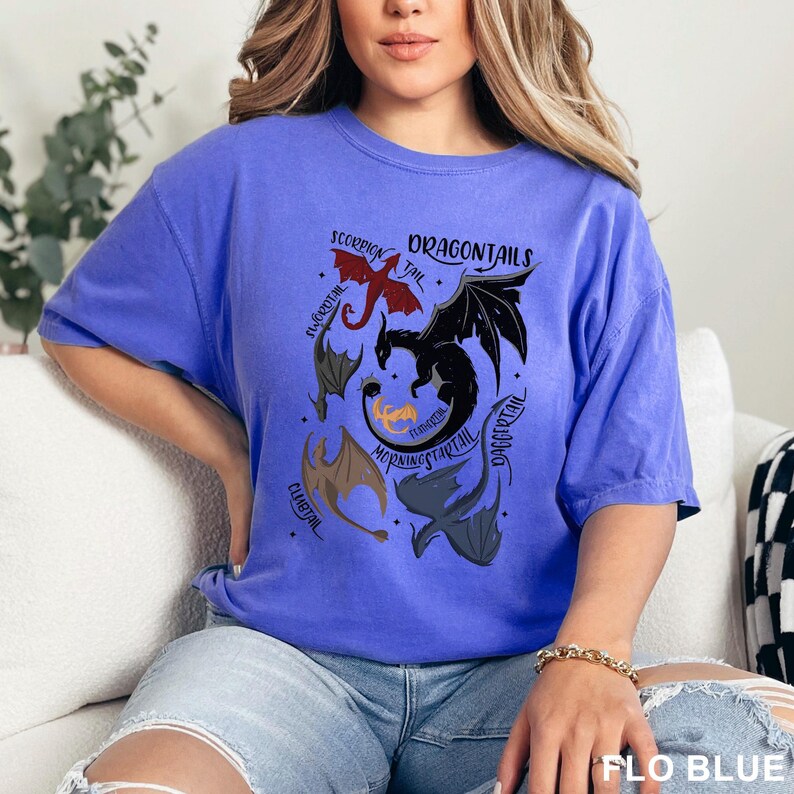以下が含まれることがあります： 赤、黒、グレーなど、さまざまな色のドラゴンが描かれた青いTシャツ。「DRAGONTAILS」の文字が上部にあり、ドラゴンの周りには他のドラゴン関連の言葉があります。シャツの底には「FLO BLUE」と書かれています。