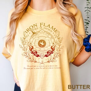 May include: Butter-colored t-shirt with a circular graphic featuring the text "IRON FLAME BURN IT DOWN." The design includes a central emblem with a bird, surrounded by flames and botanical elements. Below the graphic are two lines of text.