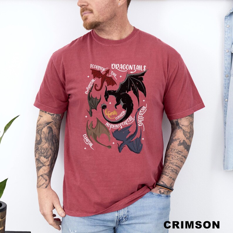 以下が含まれることがあります： 深紅色のTシャツ。ドラゴン柄と「DRAGONTAILS」の文字、ドラゴンの尻尾のイラストが描かれています。クルーネックで半袖です。「CRIMSON」の文字が下部にプリントされています。