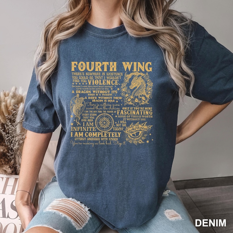 May include: A denim blue t-shirt with gold text that reads "FOURTH WING". The shirt features a dragon graphic and various phrases. The text includes "I AM INFINITE" and "I AM COMPLETELY".
