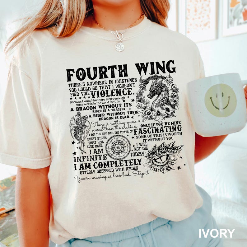 May include: Ivory-colored t-shirt with the text "Fourth Wing" and a dragon graphic. The shirt features various quotes and phrases, including "Violence" and "I am Infinite". A white mug with a smiley face is held in the image.
