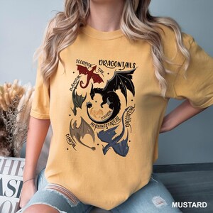 以下が含まれることがあります： マスタード色のTシャツ。赤、黒、青の様々なドラゴンテールをグラフィックデザインで表現。上部には「DRAGONTALES」の文字、ドラゴンのイラストの周りには他のテキストラベル。柔らかい素材のシャツです。