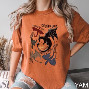 以下が含まれることがあります： オレンジ色のTシャツ。複数のドラゴンと「DRAGONTAILS」の文字が描かれたグラフィックデザイン。ドラゴンは赤、黒、青など様々な色で描かれています。Tシャツはクルーネックで半袖です。