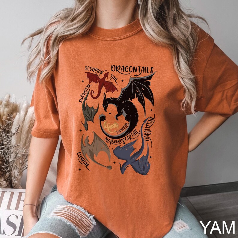 以下が含まれることがあります： オレンジ色のTシャツ。複数のドラゴンと「DRAGONTAILS」の文字が描かれたグラフィックデザイン。ドラゴンは赤、黒、青など様々な色で描かれています。Tシャツはクルーネックで半袖です。