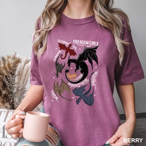 以下が含まれることがあります： ベリー色のTシャツ。デザインには「DRAGONTAILS」の文字と、さまざまなドラゴンのイラストが描かれています。各ドラゴンは異なる種類の尾でラベル付けされています。Tシャツはベリー色です。