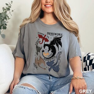 以下が含まれることがあります： ドラゴンのグラフィックが施されたグレーのTシャツ。デザインは、さまざまな色の複数のドラゴンと、「DRAGONTAILS」の文字とドラゴンの尻尾の名前が特徴です。右下隅には「GREY」の文字が印刷されています。