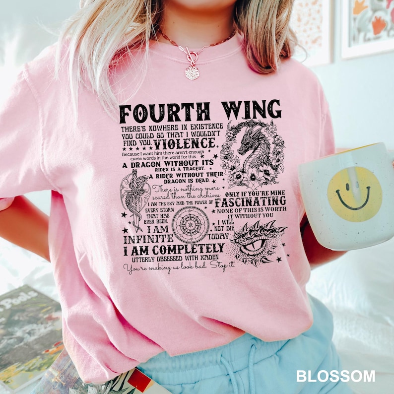May include: A light pink t-shirt with the text "FOURTH WING" and other text and graphics. The shirt also has a graphic of a dragon and a compass. A white mug with a yellow smiley face is held in the image.