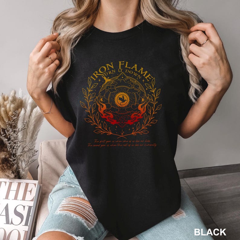 May include: Black t-shirt with a circular graphic featuring the text "IRON FLAME TURN IT DOWN" in gold. The design includes flames, a bird, and botanical elements. The shirt also has additional text at the bottom. The word "BLACK" is printed at the bottom right.