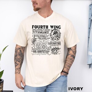 May include: Ivory-colored t-shirt with the text "FOURTH WING" and various phrases and graphics. The design includes a dragon, compass, and eye. The shirt is a crew neck and has short sleeves.