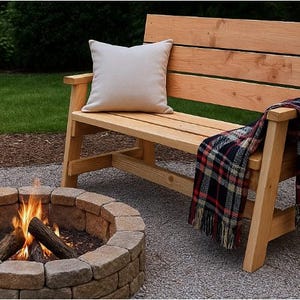 May include: A wooden outdoor bench with a light-colored cushion and a plaid blanket draped over the arm. A stone fire pit with a burning fire is in the foreground. The bench is made of light-colored wood.
