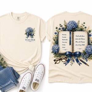 May include: A cream-colored t-shirt featuring a small blue hydrangea and bow graphic with the word TEACHER on the front. The back displays an open book with a weekly schedule, blue hydrangeas, a blue bow, eyeglasses, and a pen.
