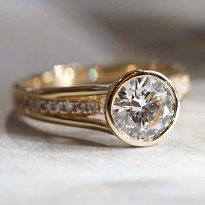 May include: A gold ring with a large, round, clear diamond center stone. The band is also gold, with smaller diamonds set around the band. The ring is set against a soft, neutral background.