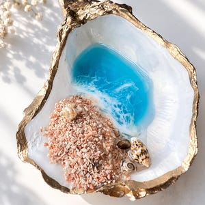 May include: A decorative oyster shell dish with a gold-painted rim. The interior features a blue resin ocean design with white foam detailing, a cluster of small pinkish-tan crushed stones, and three small natural seashells resting on the surface.