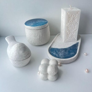 May include: A collection of white ceramic home decor items. Includes a small jar with a blue lid, a decorative candle, a tray with a blue accent, a bubble candle, and a small vase. The items are arranged on a white surface.