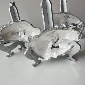 Könnte beinhalten: Silberne Austernschalen-Platzkartenhalter mit den Zahlen 2 und 5. Jede Schale wird von einem silbernen Metallsockel mit drei Füßen getragen. Hohe, silberne Metallakzente erheben sich aus den Schalen.