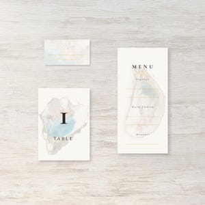 May include: A set of elegant wedding stationery featuring a watercolor design with blue, gold, and white accents. Includes a menu, place card, and table card with the number "I". The menu card has the word "MENU" at the top.