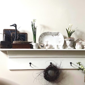 Könnte beinhalten: Ein weißes Regal mit dekorativen Gegenständen. Links befinden sich eine schwarze Holzvogelskulptur, eine kleine Holzkiste und ein gerahmtes Stück. Ein weißer Keramik-Hase und florale Teller sind ebenfalls vorhanden. Ein geflochtener Korb mit Grün hängt darunter.