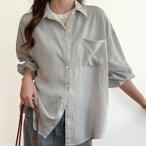 Puede incluir: Una camisa gris claro con botones y un bolsillo en el pecho. La camisa tiene mangas largas y se combina con vaqueros azules. La modelo también lleva un collar y una pulsera dorados, y lleva un bolso tote marrón.