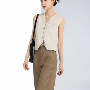 May include: A beige sleeveless vest with a V-neck and wooden buttons, paired with brown trousers. The vest has a pocket on the chest. The trousers are a solid color. The outfit is a neutral color.