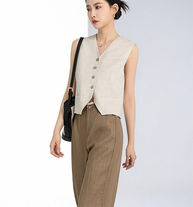 May include: A beige sleeveless vest with a V-neck and wooden buttons, paired with brown trousers. The vest has a pocket on the chest. The trousers are a solid color. The outfit is a neutral color.