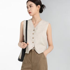 May include: A sleeveless, beige vest with a V-neck and wooden buttons. The vest is paired with brown trousers. A black shoulder bag is visible. The outfit is simple and elegant, suitable for various occasions.