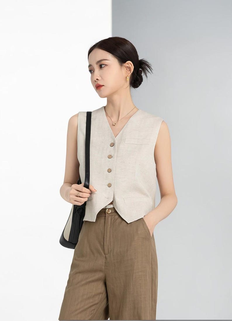 May include: A sleeveless, beige vest with a V-neck and wooden buttons. The vest is paired with brown trousers. A black shoulder bag is visible. The outfit is simple and elegant, suitable for various occasions.