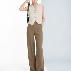 May include: A woman wearing a beige sleeveless vest with a V-neck and button closure, paired with brown wide-leg pants. The outfit is completed with sandals and a black shoulder bag. The ensemble is a neutral color palette.
