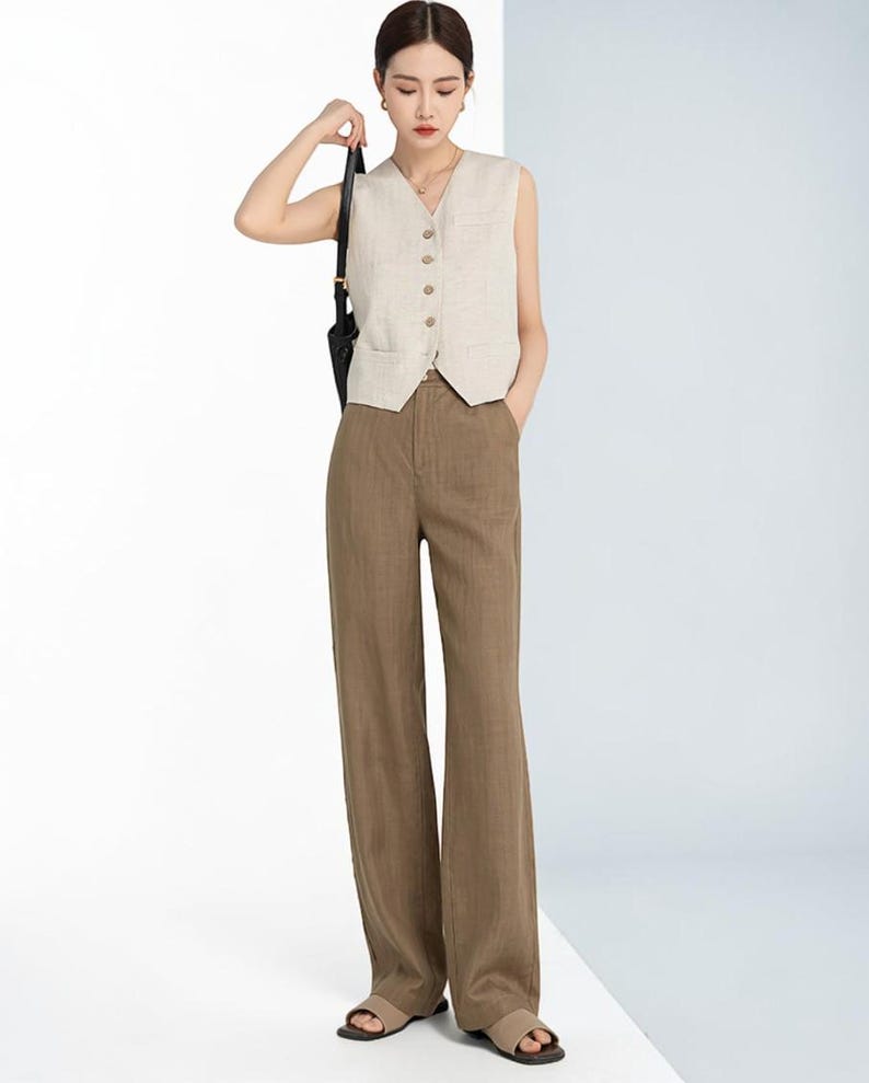 May include: A woman wearing a beige sleeveless vest with a V-neck and button closure, paired with brown wide-leg pants. The outfit is completed with sandals and a black shoulder bag. The ensemble is a neutral color palette.
