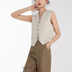 May include: A beige sleeveless vest with a V-neck, button closure, and two front pockets. The vest is paired with brown trousers. The outfit is made of a light-colored fabric, suggesting a casual or business casual style.